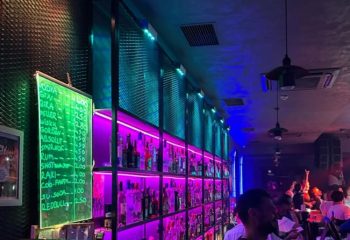 Stay Club Istanbul bar: illuminated shelves display bottles. A green chalkboard shows drink prices. Gay nightlife scene, vibrant atmosphere.