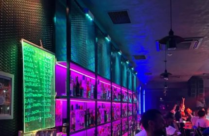 Stay Club Istanbul bar: illuminated shelves display bottles. A green chalkboard shows drink prices. Gay nightlife scene, vibrant atmosphere.