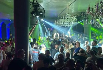 Stay Club Istanbul: A crowded gay club scene with men dancing under chandeliers and vibrant green & blue lights. Gay nightlife travel.
