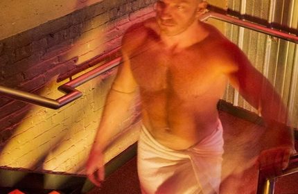 Man in towel at Steamworks bathhouse, a Chicago gay cruise club. He's on stairs, holding the rail. Red and yellow lighting.
