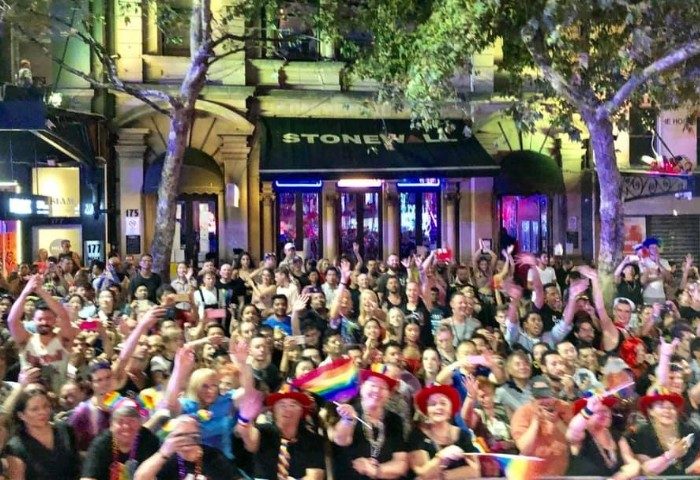 Crowd at Stonewall Hotel, Sydney. People celebrating, waving rainbow flags. Gay travel hotspot, popular LGBTQ+ venue. Night scene.