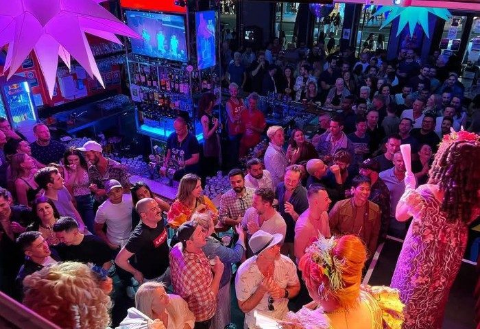 Crowded Stonewall Hotel Sydney bar. Gay men enjoy drag show, drinks, & lively atmosphere. LGBTQ+ travel destination. Vibrant nightlife scene.