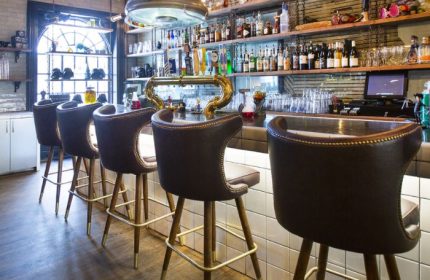 Storm Crow Manor bar in Toronto. Leather bar stools line the counter. Shelves are stocked with liquor bottles. Gay travel advice.