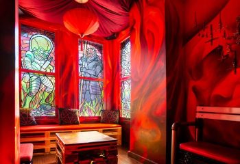 Storm Crow Manor in Toronto, Canada. Red themed room with stained glass windows, pentagram pillows, and a wooden pallet table.