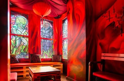 Storm Crow Manor in Toronto, Canada. Red themed room with stained glass windows, pentagram pillows, and a wooden pallet table.