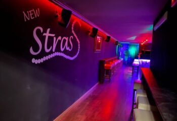 Inside Strass Barcelona gay bar: A long, narrow space with bar stools, red and purple lighting, and the Strass logo on the wall.