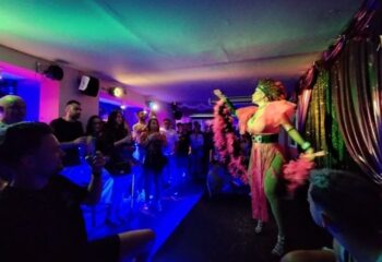 Strass Barcelona Gay Bar: Drag queen performing on stage with a pink boa in a lively gay bar. Crowd enjoying the show. Barcelona nightlife.