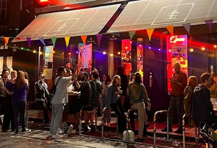 Night scene at Street 66 gay bar in Dublin. Crowds gather outside the bar, illuminated by colorful lights and rainbow flags. Gay travel.