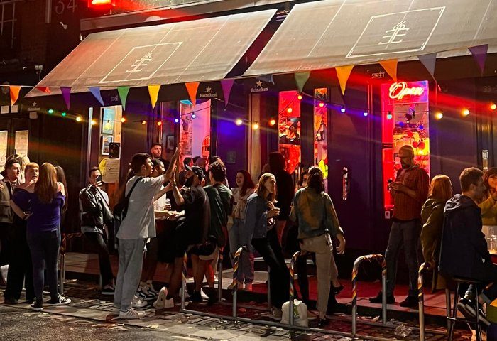 Night scene at Street 66 gay bar in Dublin. Crowds gather outside the bar, illuminated by colorful lights and rainbow flags. Gay travel.