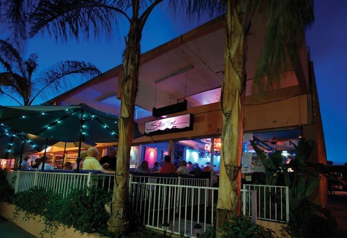 Streetbar night life: Outdoor seating at a gay bar with palm trees. People are enjoying drinks under umbrellas at this popular gay venue.