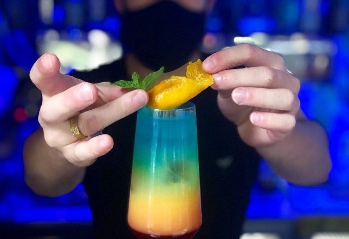 Bartender garnishing a colorful cocktail in Milan's Sui Generis gay bar. The drink features layers of red, orange, green, and blue. Mint sprig and orange peel.
