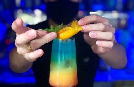 Bartender garnishing a colorful cocktail in Milan's Sui Generis gay bar. The drink features layers of red, orange, green, and blue. Mint sprig and orange peel.
