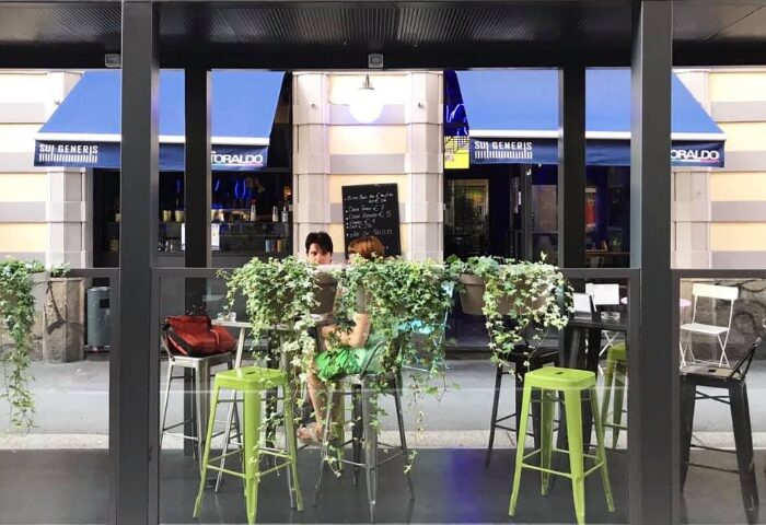 Sui Generis bar in Milan, Italy. Outdoor seating area, a popular gay-friendly spot. Blue awnings & lush greenery. Gay travel advice.