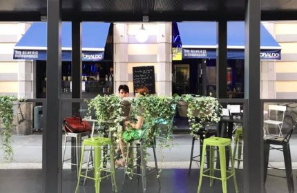 Sui Generis bar in Milan, Italy. Outdoor seating area, a popular gay-friendly spot. Blue awnings & lush greenery. Gay travel advice.