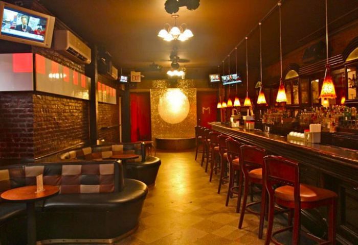 NYC gay bar interior. Lounge seating, bar stools line the counter. TVs, warm lighting, & a decorative moon backdrop create a cozy pub vibe.
