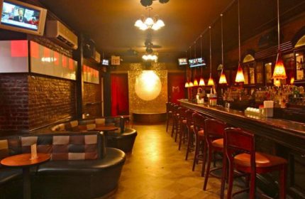 NYC gay bar interior. Lounge seating, bar stools line the counter. TVs, warm lighting, & a decorative moon backdrop create a cozy pub vibe.