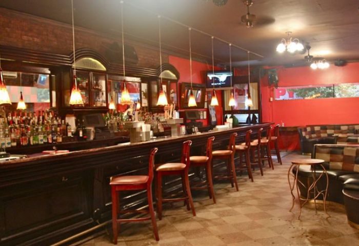NYC gay pub bar. Long bar with stools, shelves of liquor, and pendant lights. Red walls, checkered floor, and lounge seating.