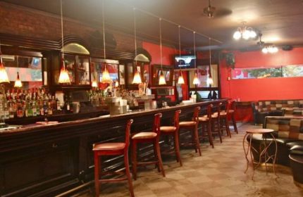 NYC gay pub bar. Long bar with stools, shelves of liquor, and pendant lights. Red walls, checkered floor, and lounge seating.