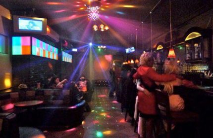 Inside Suite, a gay bar/pub in NYC. Couples dance under disco lights. TVs & colorful panels adorn the walls. A vibrant nightlife scene.