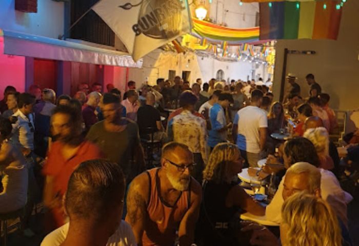 Busy gay bar in Ibiza at night, rainbow flags overhead. Gay men and women socialize at tables. Sunrise Ibiza gay bar scene.
