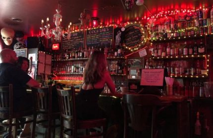 Sweaty Betty's Bar in Toronto. People at the bar, shelves of bottles, and red lights. A unique bar for gay men's travel adventures.