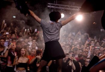 Barcelona Gay Club TANGA Party: Crowd cheers performer in striped shirt, skirt, and beret with arms raised on stage. Phones record the event.