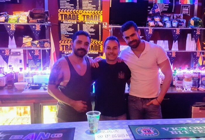Three gay men pose at TRADE Club Sydney bar. Snacks, drinks, Australian flags, and Bar Open sign visible. Gay travel advice.