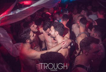 TROUGH London gay dance club scene. Shirtless men in harnesses embrace in a dark room. Nightlife at a popular gay venue.