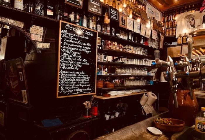 Interior of Taberna de Ángel Sierra, a gay bar in Madrid. Menu board, shelves of bottles, and bar details visible. Gay travel.