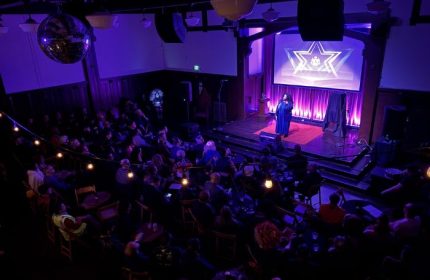 The Academy Social Club in San Francisco: Drag queen performing on stage for a gay audience. Purple lighting, disco ball, & cabaret vibe.