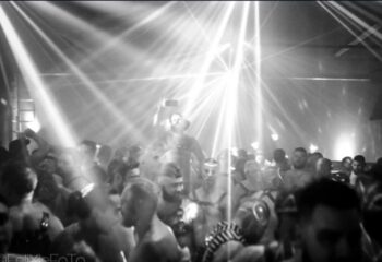 The Barracks party: Gay men in harnesses and leather gear dance at a club. Lights shine on the crowd. Black and white photo.