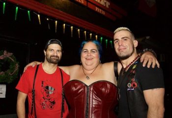 Three people at The Bullet Bar, a gay bar. Two men flank a woman in a red corset. One man wears a harness. Gay travel advice.