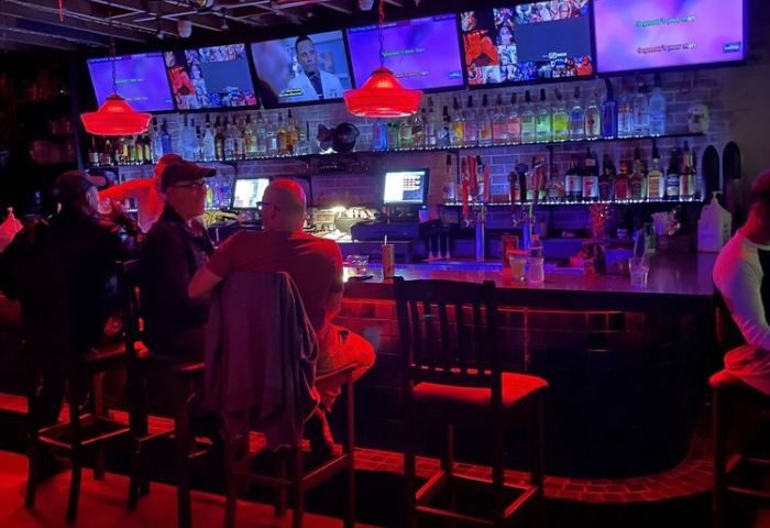 The Bullet Bar: Gay bar interior with patrons at the bar, TVs showing various content, and red lighting. Nightlife spot for gay men travelers.