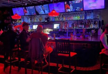 The Bullet Bar: Gay bar interior with patrons at the bar, TVs showing various content, and red lighting. Nightlife spot for gay men travelers.