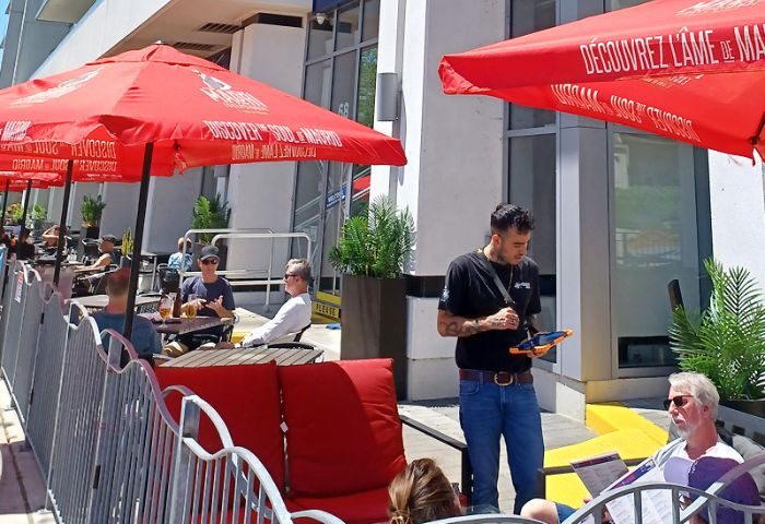 Outdoor patio at The Church Mouse Pub in Toronto. Waiter taking orders, patrons seated under red umbrellas. Gay travel.