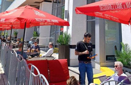 Outdoor patio at The Church Mouse Pub in Toronto. Waiter taking orders, patrons seated under red umbrellas. Gay travel.