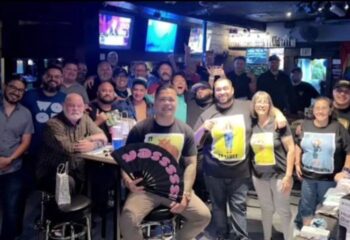 Group at The Crest Bar, a gay-friendly travel destination. People pose, some holding fans and posters, in a lively bar setting.