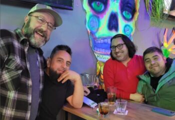 Gay men enjoying The Crest Experience at a bar. Four friends pose with drinks under a neon skull. Gay travel and nightlife scene.