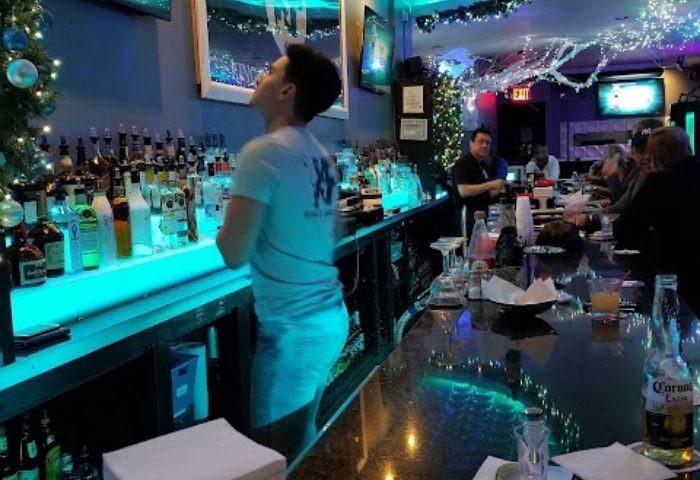 The Deep End Resto-Bar in New York: Bartender at the bar with blue lighting, bottles, and Christmas decorations. Gay bar for travel advice.