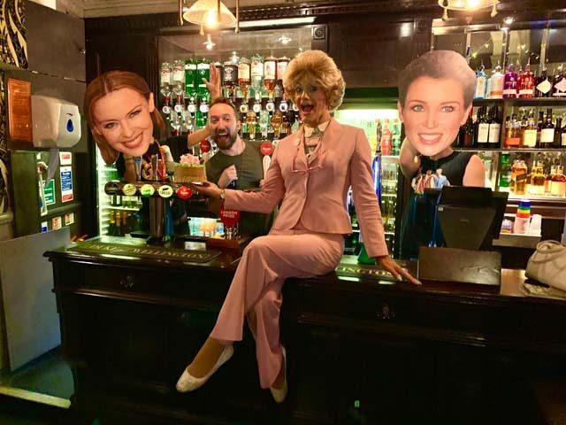 The Duke of Wellington gay bar, London. Drag queen in pink suit sits on bar, with Kylie Minogue & Lorraine Kelly cutouts. Man holding cake.