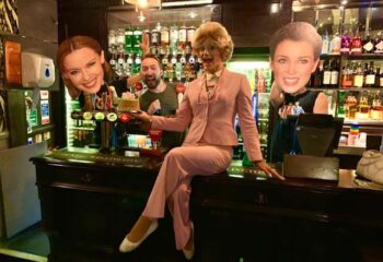The Duke of Wellington gay bar, London. Drag queen in pink suit sits on bar, with Kylie Minogue & Lorraine Kelly cutouts. Man holding cake.