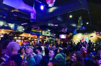 Crowded interior of The Fountainhead Pub in Vancouver. Gay bar with rainbow flags, people mingling, and a vibrant atmosphere. Gay travel advice.