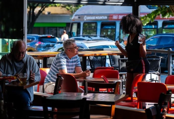 The Fountainhead Pub in Vancouver: Gay men travel. A server attends to patrons at an outdoor table. A bus is visible in the background.