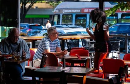 The Fountainhead Pub in Vancouver: Gay men travel. A server attends to patrons at an outdoor table. A bus is visible in the background.