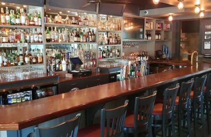 Interior view of The Glenwood gay bar in Chicago. Bar stools line the counter, liquor bottles fill shelves behind the bar.