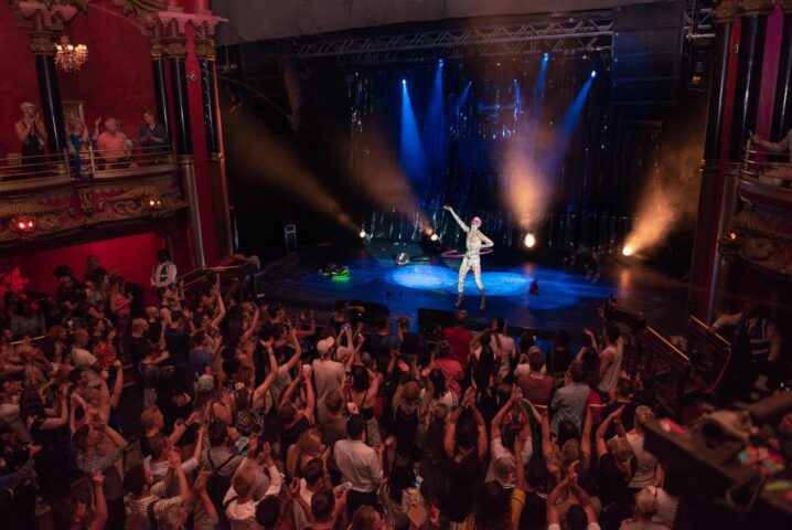 The Glory gay bar: a performer on stage with hula hoop, bathed in light, entertains a crowd in a classic theater. Gay travel advice.