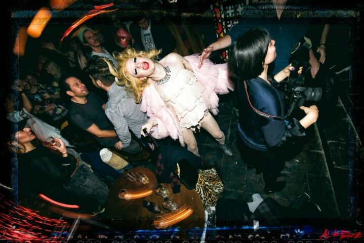 Inside The Glory gay bar: A drag queen in a pink boa & white dress is photographed in a crowded room. Gay travel nightlife scene.