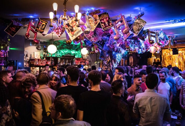 Crowded interior of The Glory Gay Bar. People gather under a ceiling adorned with posters and lights. A vibrant, lively atmosphere.