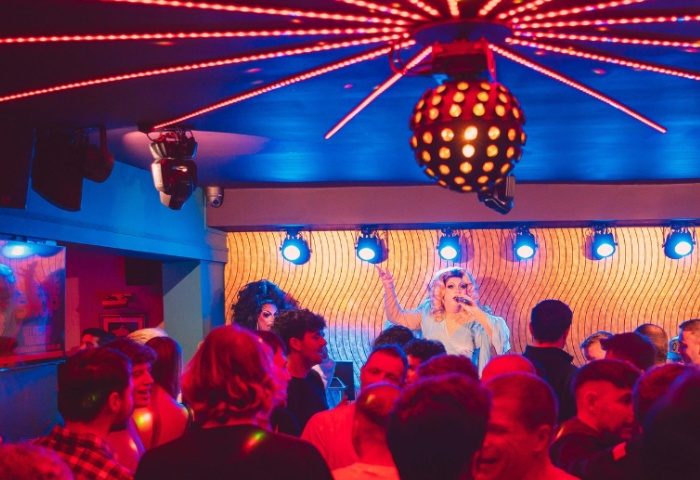 Drag queen performing at The Goose gay bar in Manchester. Crowd of gay men watching the show under disco lights. Gay travel advice.