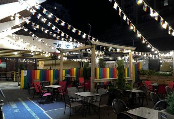 The Junction Pub in Vancouver at night. Outdoor seating area with rainbow flags and colorful decor. Gay bar travel destination.